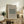 May include: Framed beige and green botanical pattern poster displayed on rustic wooden sideboard in neutral living room setting, surrounded by fern plant, books, ceramic vase, and cream upholstered chair with brown leather cushion
