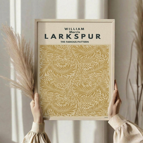 May include: Hands holding a framed botanical art print featuring William Morris's Larkspur pattern in golden yellow tones. The ornate floral design shows intricate leaves, stems and stylized flowers against cream background. Natural wood frame, displayed with dried pampas grass in soft, neutral-toned room setting with flowing curtains.
