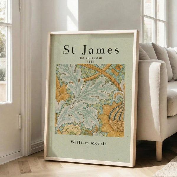 May include: Framed art print with sage green background featuring ornate botanical pattern in golden yellow and mint green tones, displaying "St James The MET Museum 1881 William Morris" text. Light wood frame leaning against white wall beside beige upholstered chair in bright, naturally-lit room with hardwood floors.