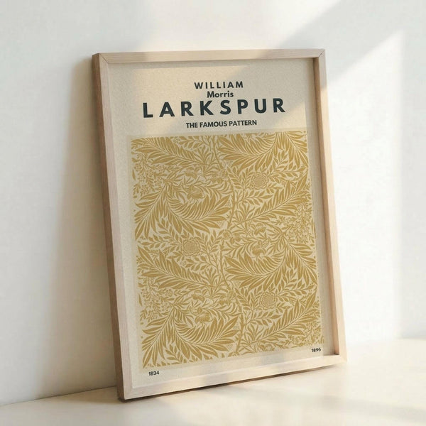 Framed botanical print featuring intricate golden yellow foliage pattern on cream background. The detailed leaf and branch design fills the center area, with "WILLIAM Morris LARKSPUR THE FAMOUS PATTERN" text at top and "1834" and "1896" dates in corners. Light wood frame sits propped against white wall on white surface, casting subtle shadows.
