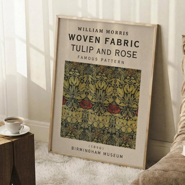 May include: Framed poster featuring William Morris's Tulip and Rose woven fabric pattern from 1890, displayed in a cozy room setting. The artwork shows an intricate floral design in muted greens and yellows with red rose accents on a beige background. The poster leans against white paneled walls next to a wooden block side table with a white teacup and saucer, beside a beige knit throw blanket on a textured cream rug.