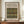 May include: Framed botanical poster displaying William Morris's Tulip and Rose fabric pattern in green, gold, and red tones on cream background, positioned on wooden sideboard beside white ceramic vase and stacked books against neutral wall with white molding