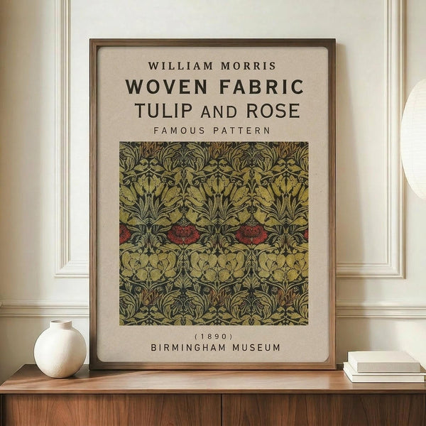 May include: Framed botanical poster displaying William Morris's Tulip and Rose fabric pattern in green, gold, and red tones on cream background, positioned on wooden sideboard beside white ceramic vase and stacked books against neutral wall with white molding