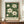 May include: Framed botanical poster with white magnolia flowers and green leaves against dark emerald background, displayed on wooden sideboard next to white ceramic vase and stacked books in elegant room with cream walls and white molding