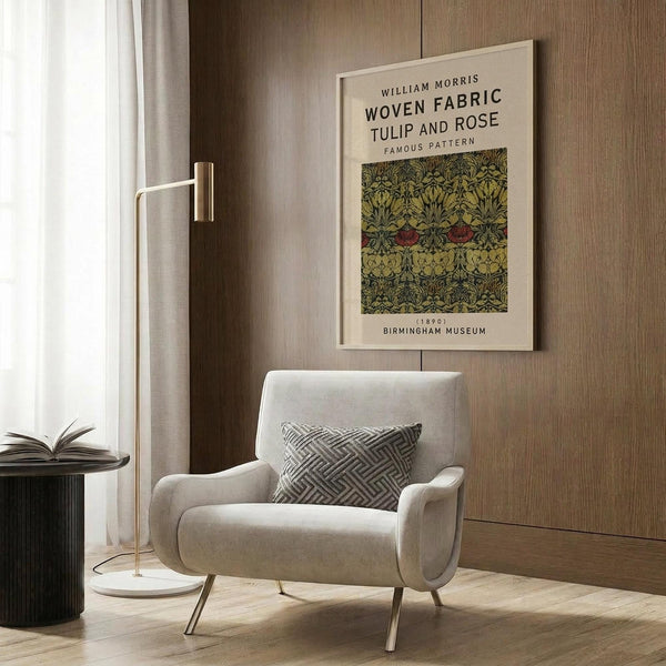 May include: Cream-colored framed poster featuring William Morris's Tulip and Rose fabric pattern in green and burgundy tones, displayed on wood paneled wall above beige upholstered armchair with geometric patterned pillow, brass floor lamp with white marble base nearby, creating warm modern interior setting with natural lighting from window.