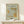 May include: A framed poster with ornate botanical design in sage green and golden orange tones featuring flowing acanthus leaves and decorative swirls, displayed on a light wood console table beside a small white wrapped gift with natural twine and evergreen sprig against a cream wall.