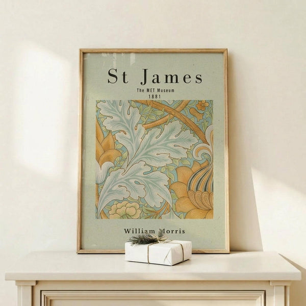 May include: A framed poster with ornate botanical design in sage green and golden orange tones featuring flowing acanthus leaves and decorative swirls, displayed on a light wood console table beside a small white wrapped gift with natural twine and evergreen sprig against a cream wall.