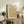 May include: Framed botanical poster with golden yellow leaf pattern displayed on rustic wooden sideboard next to cream upholstered chair, fern plant, ceramic vases and books. The print features intricate flowing foliage design in warm mustard tones with black text header against cream background in natural wood frame.