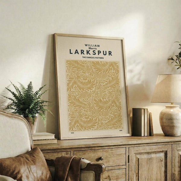 May include: Framed botanical poster with golden yellow leaf pattern displayed on rustic wooden sideboard next to cream upholstered chair, fern plant, ceramic vases and books. The print features intricate flowing foliage design in warm mustard tones with black text header against cream background in natural wood frame.