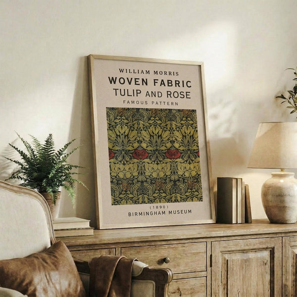 May include: Framed vintage botanical textile pattern poster displayed on rustic wooden sideboard in neutral living room. Shows intricate green and burgundy floral design with tulips and roses. Light wood frame, surrounded by fern plant, ceramic vase, stacked books, and beige upholstered furniture. Warm natural lighting creates cozy interior styling scene.
