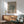 May include: Framed William Morris cotton pattern poster in sage green and cream tones hanging above mid-century modern wooden sideboard. Scene shows mint green upholstered armchair with wooden frame, small round side table, and white ceramic table lamp. Warm natural sunlight creates geometric shadows across white wall. Poster features vintage textile design with "Cotton" and "Eye Bright" text in minimalist typography within thin copper-toned frame.