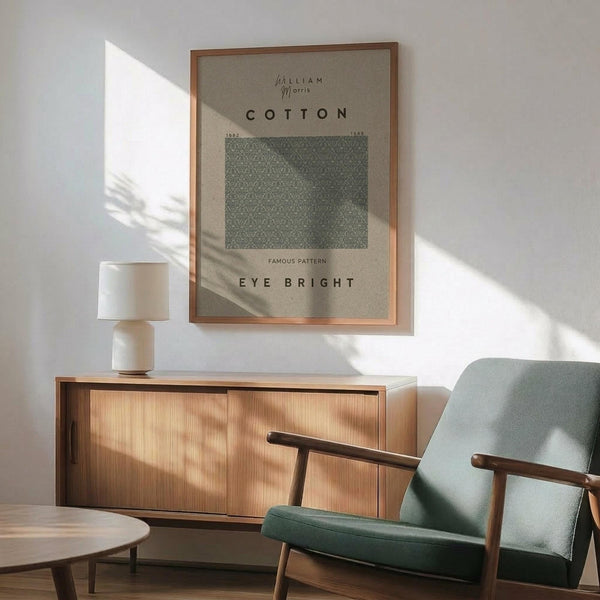 May include: Framed William Morris cotton pattern poster in sage green and cream tones hanging above mid-century modern wooden sideboard. Scene shows mint green upholstered armchair with wooden frame, small round side table, and white ceramic table lamp. Warm natural sunlight creates geometric shadows across white wall. Poster features vintage textile design with "Cotton" and "Eye Bright" text in minimalist typography within thin copper-toned frame.
