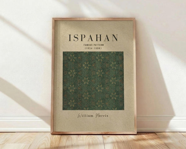 May include: Framed poster with beige background displaying green ornate floral pattern sample, black text reading "ISPAHAN FAMOUS PATTERN (1834-1896)" and "William Morris" signature, natural wood frame leaning against white wall on light wooden floor with soft window shadows