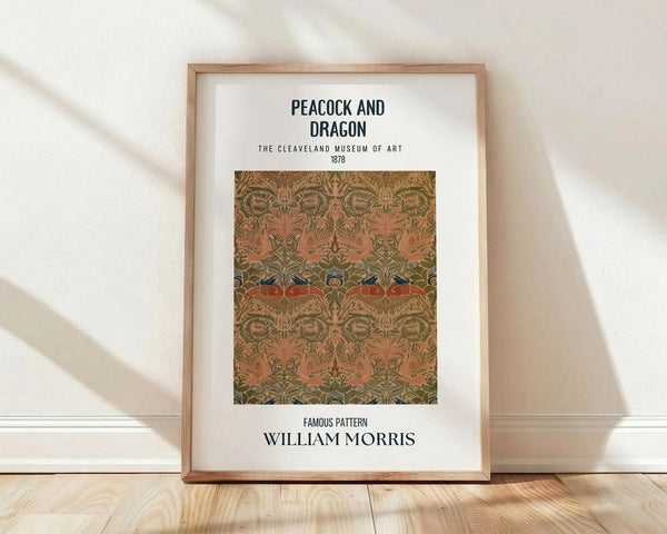 May include: Framed poster print featuring William Morris's ornate Peacock and Dragon pattern from 1878 in earthy tones of coral, olive green, and gold. The symmetrical design shows stylized peacocks and mythical creatures amid flowing botanical motifs. Natural wood frame with white matting, displayed on light hardwood floor against white wall with soft natural lighting creating diagonal shadows.