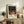 May include: Framed vintage exhibition poster featuring orange deer in whimsical forest scene with white dotted night sky and red moons, displayed on rustic wooden sideboard alongside fern plant, ceramic vase, and books in neutral-toned living room setting with beige armchair