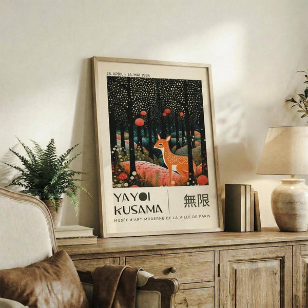 May include: Framed vintage exhibition poster featuring orange deer in whimsical forest scene with white dotted night sky and red moons, displayed on rustic wooden sideboard alongside fern plant, ceramic vase, and books in neutral-toned living room setting with beige armchair