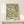May include: Framed botanical art print featuring flowing green and cream foliage pattern on beige background, displayed on light wood console table next to small wrapped gift with twine and greenery sprig against cream wall