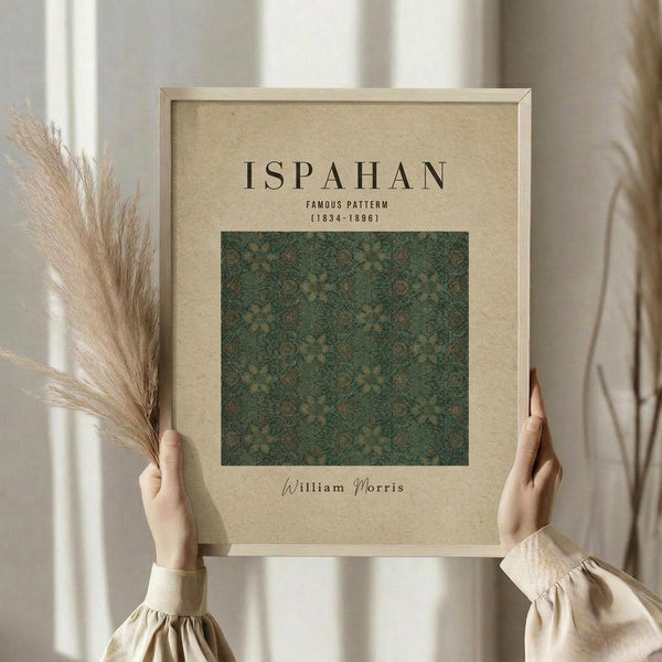 May include: Hands holding a framed art print featuring an intricate green and gold floral pattern in Victorian style. The beige matted frame displays text reading "ISPAHAN FAMOUS PATTERN (1834-1896)" and "William Morris" below the ornate botanical design. Dried pampas grass stems visible on the left side against a soft neutral background with natural window lighting.