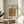 May include: Framed vintage-style poster displaying William Morris's ornate Peacock and Dragon textile pattern from 1878, featuring intricate floral motifs in coral, green, and blue tones. The cream-colored print sits on a round wooden side table next to a beige upholstered chair, with natural sunlight streaming across white walls and a plant visible in the background.