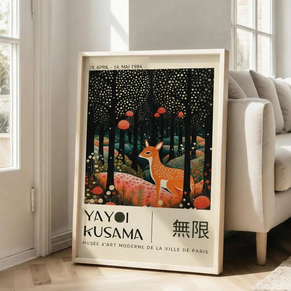 May include: Framed art print featuring an orange deer with white spots sitting in a dark forest scene with red mushrooms, scattered white dots resembling snow or stars, and tall dark trees. The poster shows exhibition text "YAYOI KUSAMA" and Japanese characters below the artwork, dated 1984, referencing Musee d'Art Moderne de la Ville de Paris. The light wood frame leans against a white wall next to a beige upholstered sofa in a bright room with large windows.