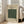 May include: Framed vintage-style poster with cream background featuring green ornate paisley pattern design, displayed leaning against white wall next to beige upholstered chair in bright living room with large windows and wooden floors