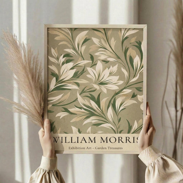 May include: Hands holding a framed botanical art print featuring flowing cream and sage green foliage patterns on an olive background. The minimalist poster displays "William Morris" and "Exhibition Art - Garden Treasures" text below the ornate leaf design. Natural wheat stems visible to the left, soft beige curtains in background, person wearing cream colored clothing with gathered sleeves.