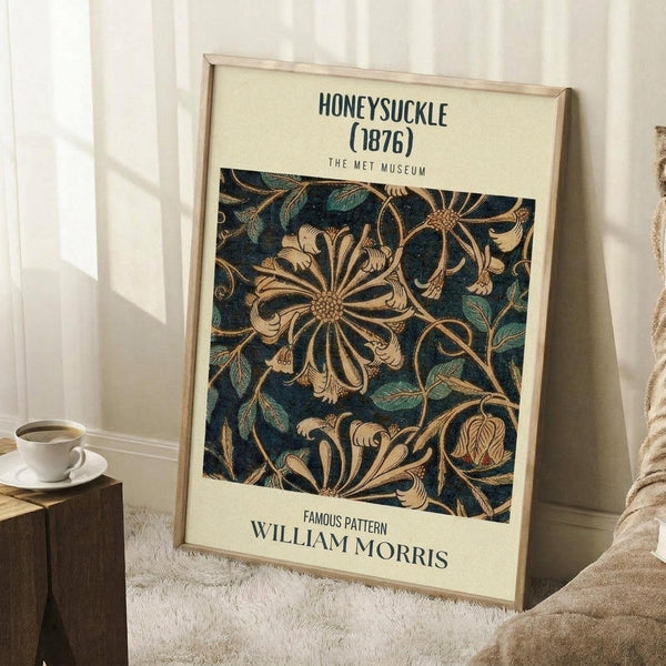 May include: Framed poster with cream background displaying ornate floral pattern in deep blue and golden tones, featuring stylized honeysuckle flowers and curved leaves in William Morris's distinctive Arts and Crafts style. Frame leans against white paneled wall next to white coffee cup on wooden surface and textured beige throw blanket on white fluffy rug.