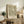 May include: Framed botanical print with flowing green and cream leaf pattern displayed on rustic wooden sideboard alongside fern plant, stacked books, and ceramic vase in neutral-toned living room setting with beige chair and soft natural lighting