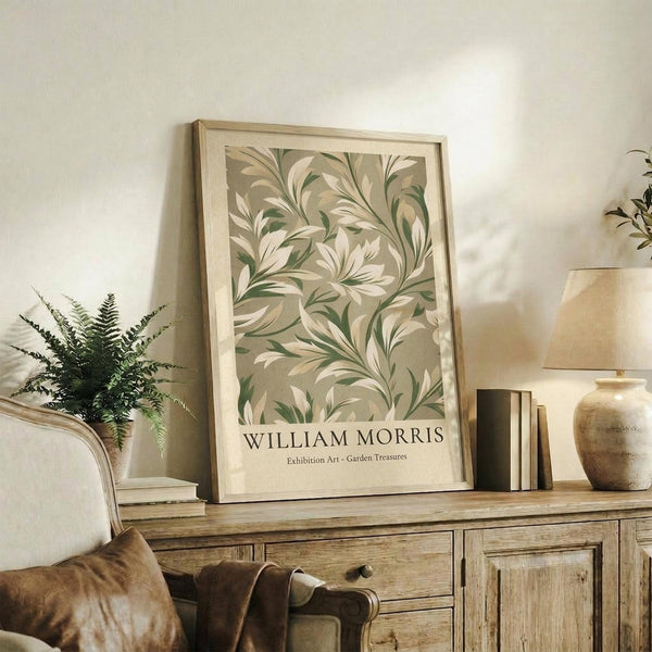 May include: Framed botanical print with flowing green and cream leaf pattern displayed on rustic wooden sideboard alongside fern plant, stacked books, and ceramic vase in neutral-toned living room setting with beige chair and soft natural lighting