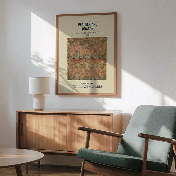 May include: Framed William Morris Peacock and Dragon pattern print hanging on white wall above wooden sideboard with ceramic table lamp. Mid-century modern sage green upholstered armchair with wooden frame sits nearby. The vintage-style poster shows ornate peacocks and foliage in muted greens, pinks and earth tones. Warm natural sunlight streams across the minimalist Scandinavian-inspired interior setting.