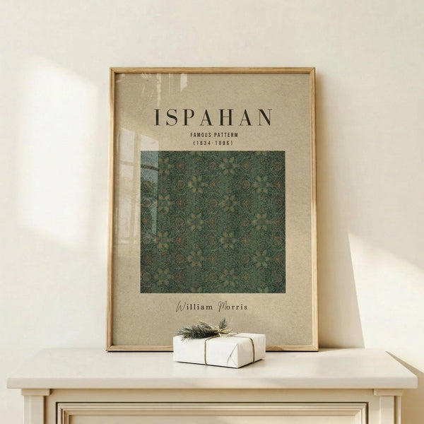 May include: Framed poster with beige background displaying "ISPAHAN FAMOUS PATTERN (1834-1896)" text above a green botanical pattern square, "William Morris" signature below. Light wood frame on white wooden table with small white wrapped gift tied with twine and greenery sprig against cream wall.