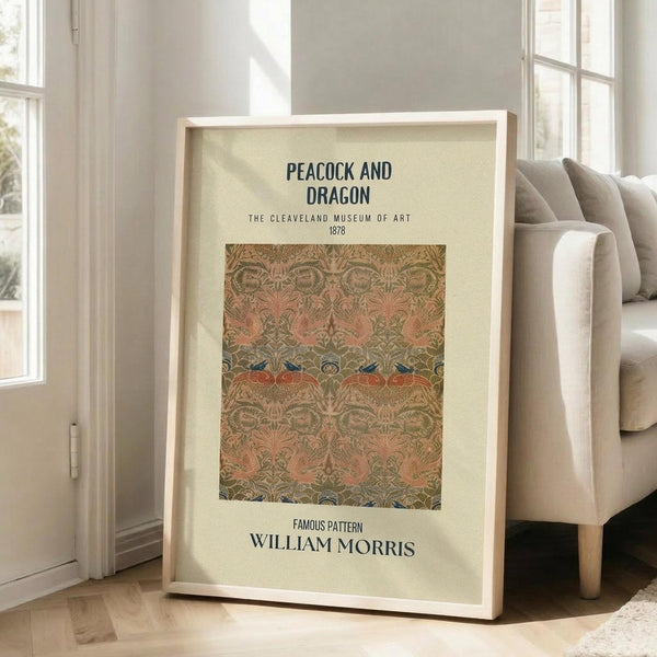 May include: Framed poster with cream background displaying ornate textile pattern in earthy tones of salmon pink, sage green, and gold. Features repeating motifs of stylized peacocks and dragons in Art Nouveau style. Black text identifies museum and artist. Frame leans against white wall next to beige upholstered chair in bright, naturally-lit room with white baseboards.
