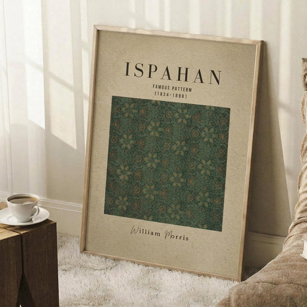 May include: Framed poster with beige background displaying "ISPAHAN FAMOUS PATTERN (1834-1898)" text above green ornate floral pattern square, "William Morris" signature below. Natural wood frame leaning against white wall corner near wooden side table with white coffee cup, beige throw blanket visible, soft natural lighting creates gentle shadows.