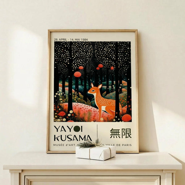 May include: Framed exhibition poster featuring a spotted deer in a whimsical forest scene with white polka dots on black sky, orange mushrooms, and colorful foliage. Text shows "YAYOI KUSAMA" and Japanese characters below the artwork. Light wood frame sits on white wooden surface next to small wrapped gift with twine and evergreen sprig against cream wall.