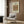 May include: Cream-colored poster with ornate peacock and dragon textile pattern in earthy tones, displayed on wood-paneled wall above modern beige armchair with geometric pillow. Scene shows contemporary reading nook with brass floor lamp and side table with open book, natural lighting from window with sheer curtains.