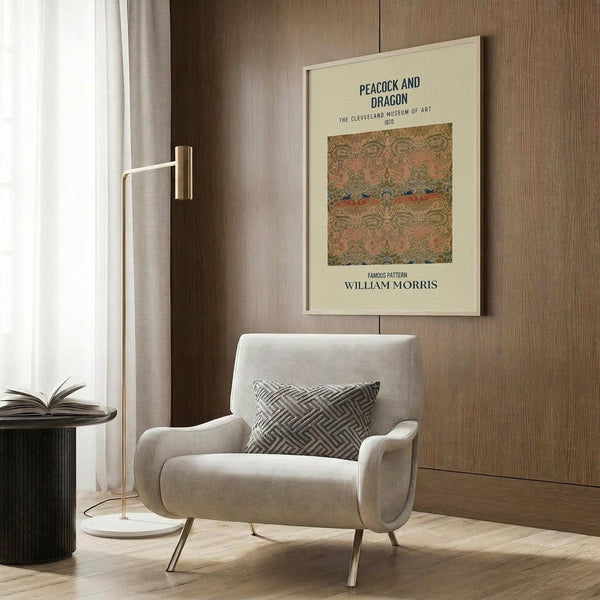 May include: Cream-colored poster with ornate peacock and dragon textile pattern in earthy tones, displayed on wood-paneled wall above modern beige armchair with geometric pillow. Scene shows contemporary reading nook with brass floor lamp and side table with open book, natural lighting from window with sheer curtains.