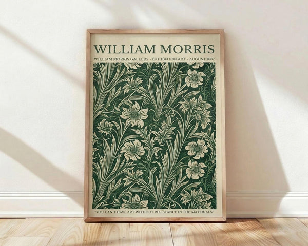 May include: Framed botanical print with dark green background featuring cream-colored flowers and flowing foliage in vintage Arts and Crafts style. Natural wood frame leans against white wall on light wooden floor with soft shadows from window light. Quote text appears at bottom of artwork.