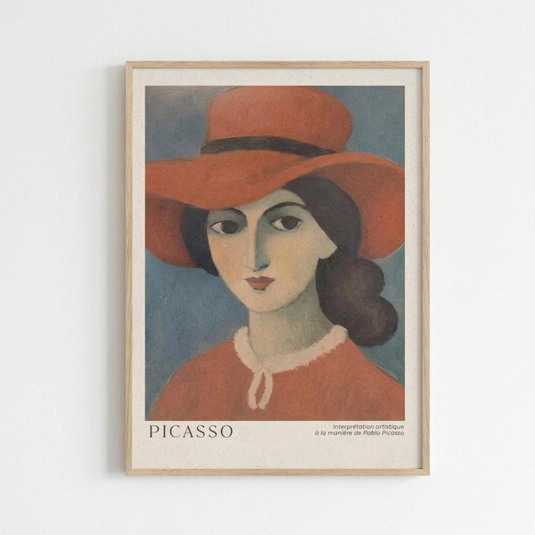 May include: Framed art print showing a woman wearing an orange-red wide-brimmed hat and matching top with white collar detail. The portrait features simplified facial features and dark hair in a modernist style reminiscent of early 20th century art. The piece has a blue-gray background and is displayed in a light wood frame with white matting against a white wall. Text at bottom reads "PICASSO" with smaller French text.