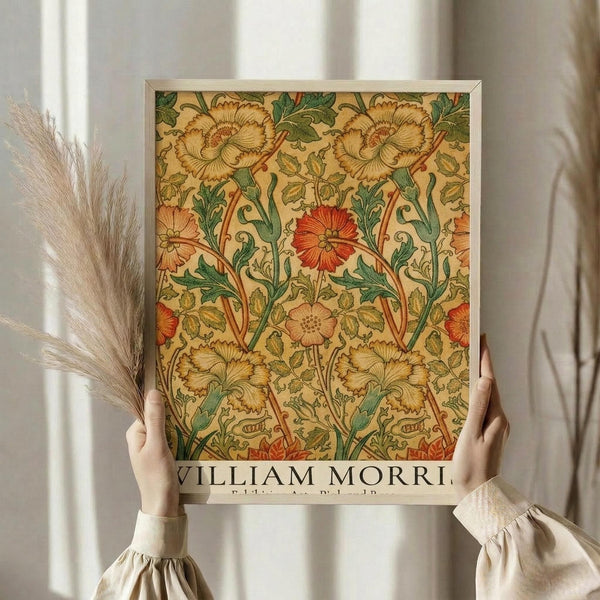 May include: Person in cream sweater holding framed botanical art print featuring William Morris floral pattern with large blooms in yellow, orange, and red tones with green foliage on cream background. White wooden frame, dried pampas grass visible to left, soft natural lighting against neutral curtained background.