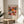 May include: Framed exhibition poster featuring colorful circular floral and dotted patterns in pink, orange, and coral tones against a dark background, displayed on a light wood round side table next to a beige upholstered chair. Sunlight creates soft shadows on the neutral wall behind, with green plant leaves visible in the corner of the minimalist room setting.