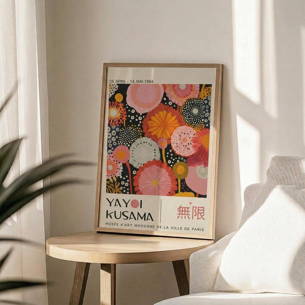 May include: Framed exhibition poster featuring colorful circular floral and dotted patterns in pink, orange, and coral tones against a dark background, displayed on a light wood round side table next to a beige upholstered chair. Sunlight creates soft shadows on the neutral wall behind, with green plant leaves visible in the corner of the minimalist room setting.