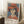 May include: Framed art print showing a woman wearing a large red-orange hat and matching dress with white collar details, painted in a modernist style with geometric facial features against a blue background. The poster displays "PICASSO" text at bottom and leans against a white wall on a textured cream rug, with a coffee cup on a wooden surface nearby and soft natural lighting.