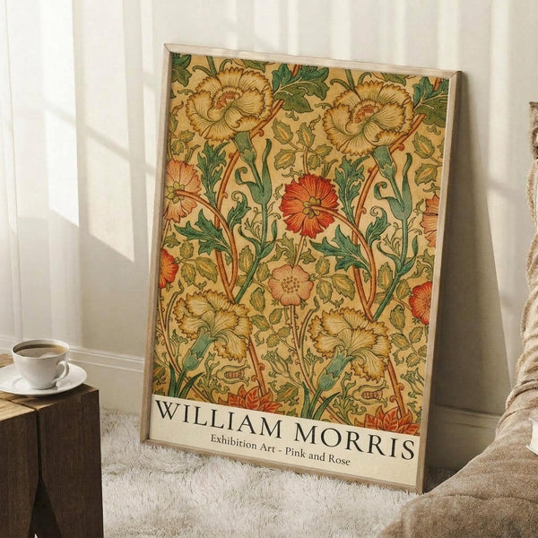 May include: Framed botanical poster with William Morris floral pattern featuring golden carnations, red poppy flowers, and green foliage on cream background, leaning against white wall beside wooden side table with white coffee cup and saucer, on textured white rug with beige throw blanket visible.