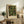 May include: Framed botanical art print featuring colorful fruit and flower pattern with green leaves, yellow lemons, orange peaches, and red berries on dark background. Light wood frame displayed on rustic wooden sideboard alongside books, ceramic vase, and fern plant in neutral-toned living room setting.
