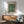 May include: Framed botanical poster with green floral pattern hanging above wooden sideboard. Mid-century modern living room setting with sage green upholstered armchair, round coffee table, and white ceramic table lamp. Natural sunlight creates shadows on white wall. Wood frame complements warm wood furniture tones.