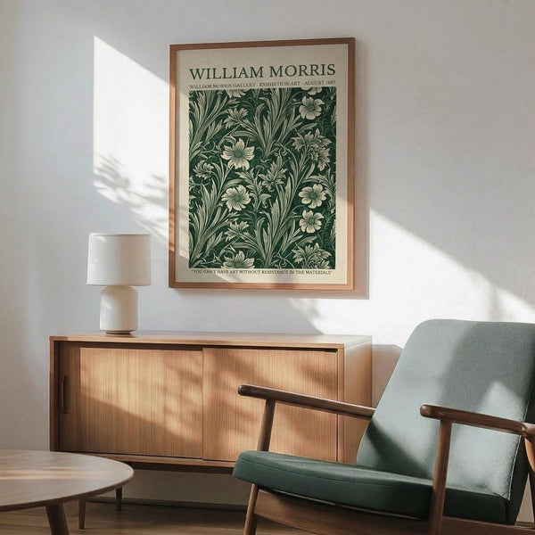 May include: Framed botanical poster with green floral pattern hanging above wooden sideboard. Mid-century modern living room setting with sage green upholstered armchair, round coffee table, and white ceramic table lamp. Natural sunlight creates shadows on white wall. Wood frame complements warm wood furniture tones.
