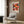 May include: Colorful Yayoi Kusama exhibition poster with vibrant circular patterns in pink, orange, and black displayed in light wooden frame on wood-paneled wall above cream upholstered armchair with geometric throw pillow. Modern brass floor lamp and dark round side table with open book complete the contemporary living room setting with natural lighting from window.
