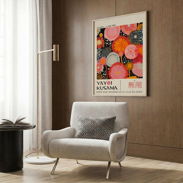 May include: Colorful Yayoi Kusama exhibition poster with vibrant circular patterns in pink, orange, and black displayed in light wooden frame on wood-paneled wall above cream upholstered armchair with geometric throw pillow. Modern brass floor lamp and dark round side table with open book complete the contemporary living room setting with natural lighting from window.