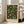 May include: Hands holding a framed botanical poster featuring William Morris's fruit pattern design with green leaves, yellow and orange fruits, and small flowers on a dark background. The print is displayed in a bright living room with white walls, large windows, and a beige sofa. The wooden frame has a light natural finish.