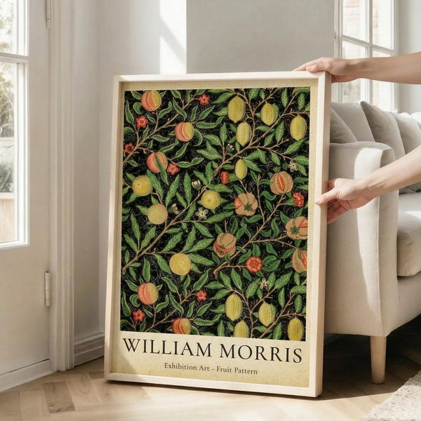 May include: Hands holding a framed botanical poster featuring William Morris's fruit pattern design with green leaves, yellow and orange fruits, and small flowers on a dark background. The print is displayed in a bright living room with white walls, large windows, and a beige sofa. The wooden frame has a light natural finish.
